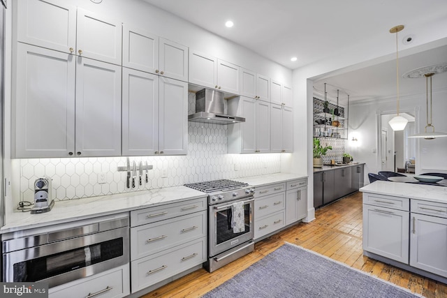 kitchen with appliances with stainless steel finishes, wall chimney exhaust hood, light wood-type flooring, backsplash, and light stone counters