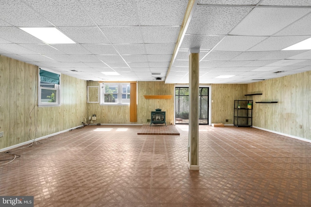 basement featuring a wood stove, wood walls, tile patterned floors, baseboards, and a paneled ceiling