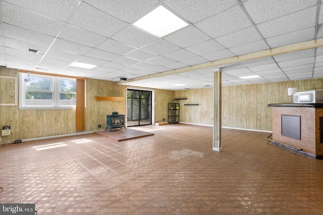 below grade area with tile patterned floors, a wood stove, a drop ceiling, wooden walls, and baseboards