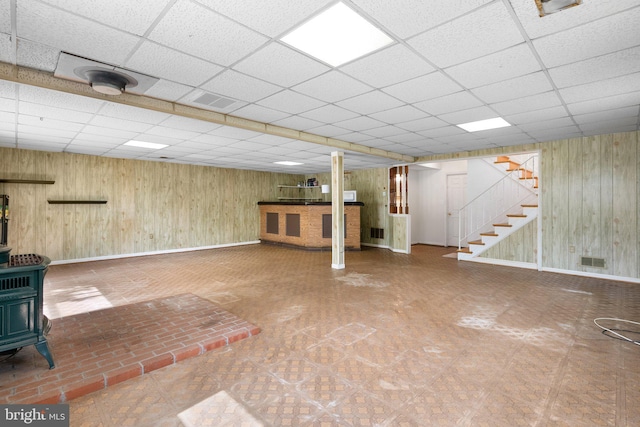 finished basement featuring a paneled ceiling, a wood stove, baseboards, stairs, and wood walls