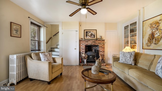 living room featuring a ceiling fan, light wood-style floors, radiator heating unit, stairway, and a wood stove