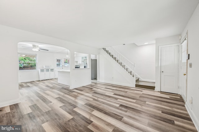 unfurnished living room with arched walkways, light wood-style floors, stairway, a ceiling fan, and baseboards