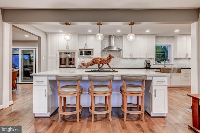 kitchen with wall chimney range hood, stainless steel appliances, white cabinetry, light wood-style floors, and recessed lighting