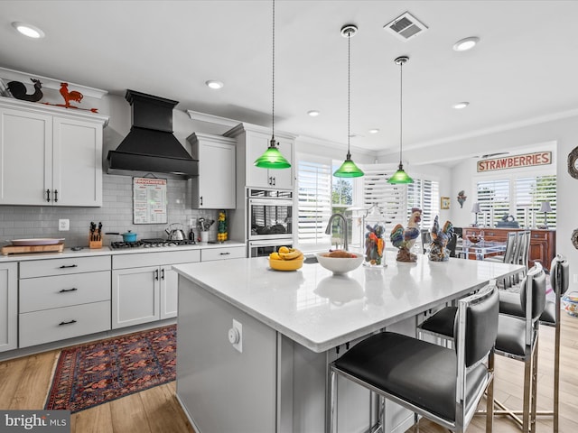 kitchen with custom exhaust hood, appliances with stainless steel finishes, tasteful backsplash, light wood finished floors, and a kitchen island with sink