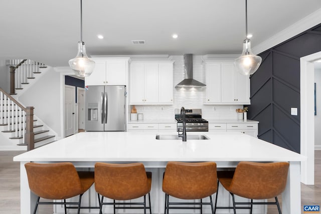 kitchen featuring appliances with stainless steel finishes, wall chimney exhaust hood, a kitchen island with sink, a sink, and white cabinetry