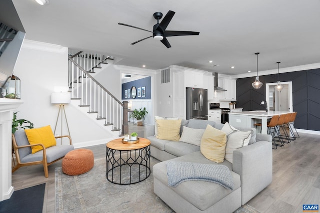 living area with light wood-style flooring, a ceiling fan, ornamental molding, baseboards, and stairs