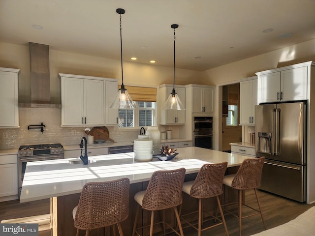 kitchen featuring appliances with stainless steel finishes, wall chimney range hood, light countertops, a kitchen island with sink, and recessed lighting