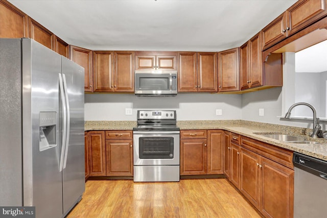 kitchen featuring stainless steel appliances, a sink, light wood-style floors, brown cabinetry, and light stone countertops
