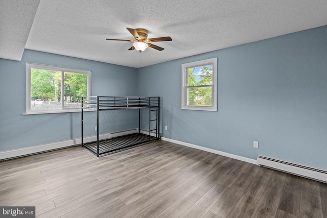 unfurnished bedroom featuring wood finished floors, a baseboard heating unit, a textured ceiling, baseboards, and a ceiling fan