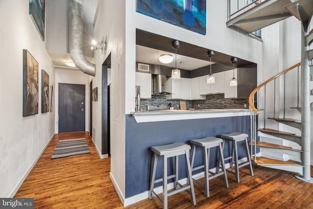 kitchen with wall chimney exhaust hood, hardwood / wood-style flooring, a kitchen breakfast bar, decorative backsplash, and a towering ceiling