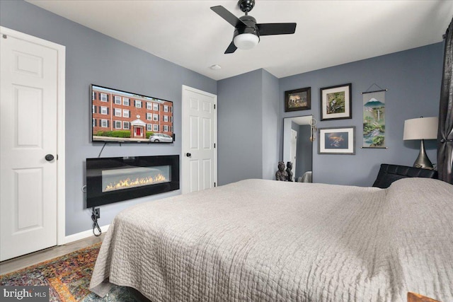 bedroom featuring wood finished floors, a glass covered fireplace, baseboards, and ceiling fan
