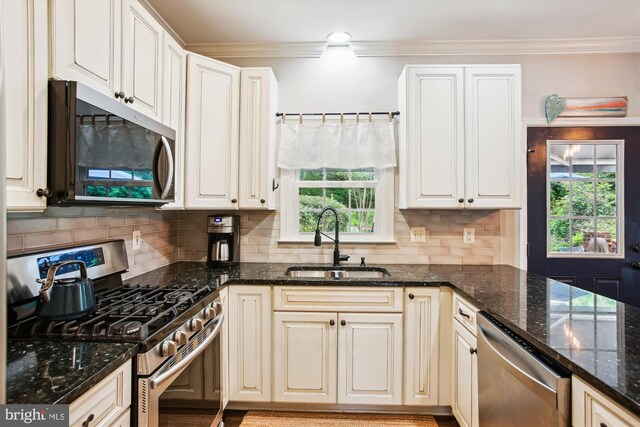 kitchen featuring appliances with stainless steel finishes, a sink, ornamental molding, plenty of natural light, and dark stone counters