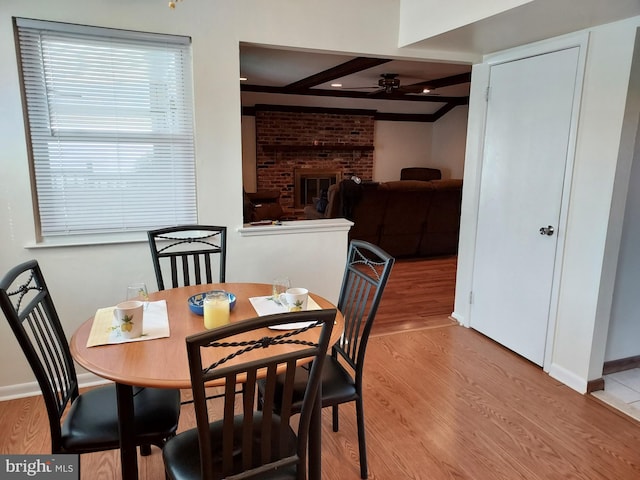 dining room featuring a ceiling fan, beam ceiling, baseboards, wood finished floors, and a brick fireplace