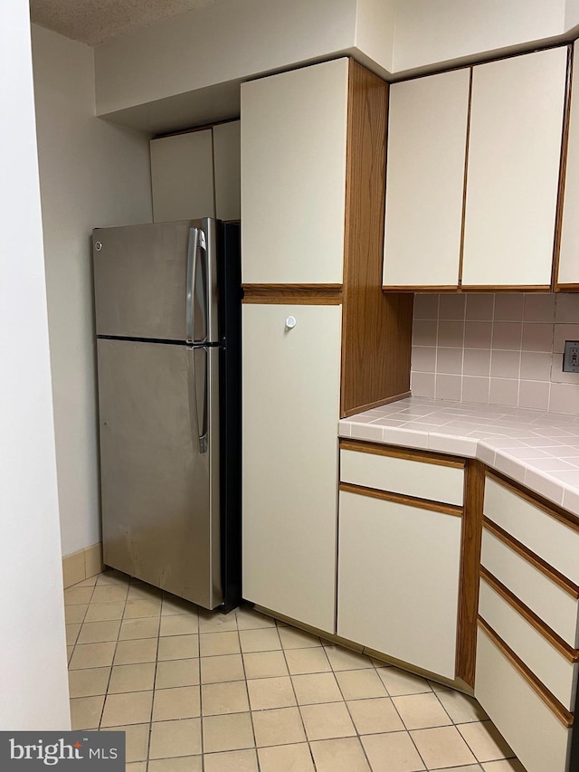 kitchen with freestanding refrigerator, tile countertops, backsplash, light tile patterned flooring, and white cabinetry