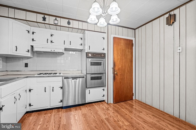 kitchen with stainless steel appliances, a chandelier, under cabinet range hood, white cabinets, and wood walls