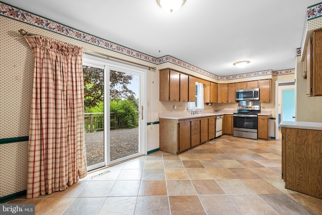 kitchen with stainless steel appliances, wallpapered walls, light countertops, brown cabinetry, and a sink