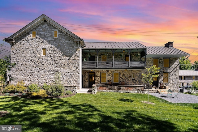 back of property at dusk with a balcony, a standing seam roof, stone siding, and a lawn