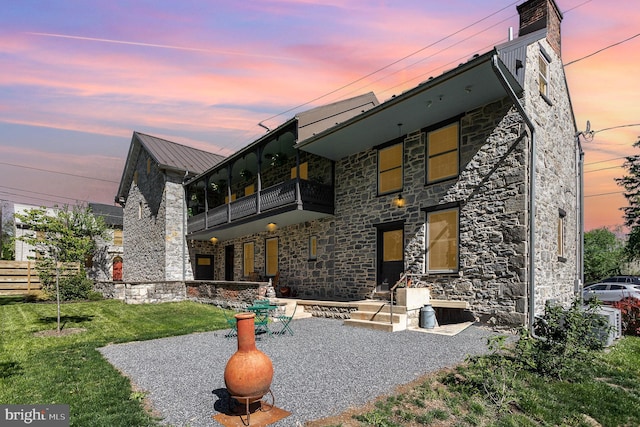 back of property featuring a balcony, stone siding, a yard, metal roof, and a patio