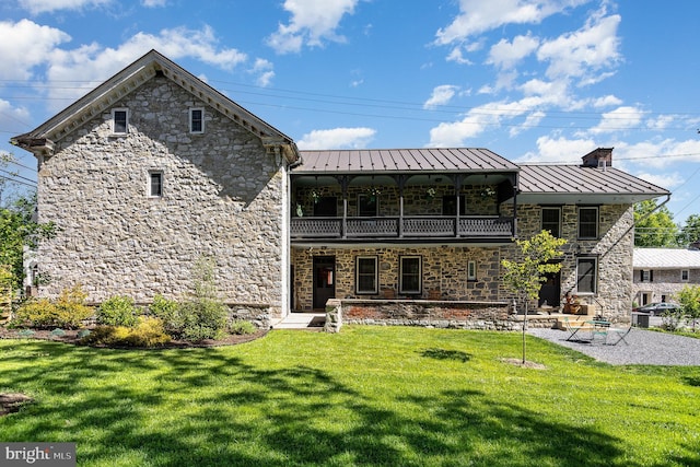 rear view of property featuring a balcony, stone siding, a standing seam roof, a yard, and a patio area