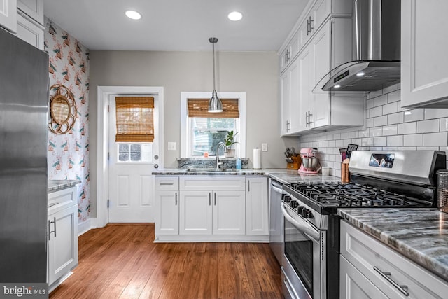kitchen with stainless steel appliances, wall chimney range hood, a sink, wood finished floors, and white cabinets