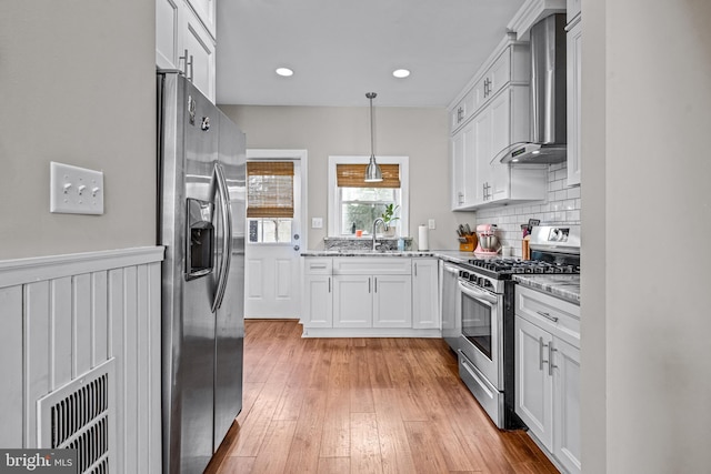 kitchen featuring appliances with stainless steel finishes, wall chimney range hood, white cabinets, light stone counters, and hanging light fixtures