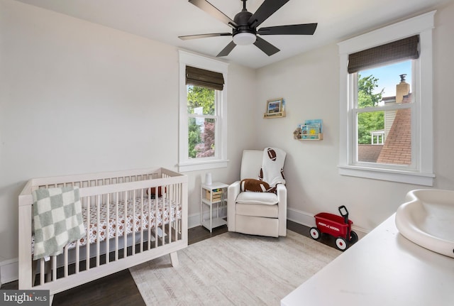 bedroom with wood finished floors, a nursery area, baseboards, and a ceiling fan