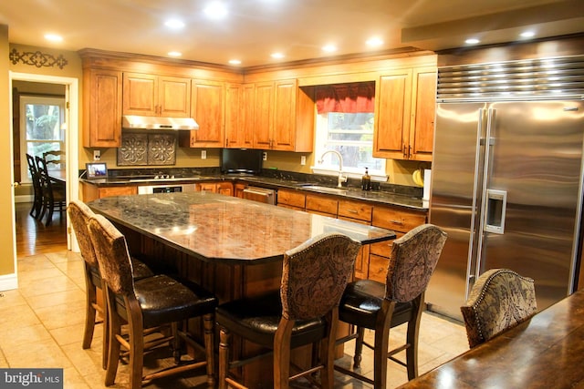 kitchen featuring appliances with stainless steel finishes, a sink, under cabinet range hood, light tile patterned flooring, and a center island