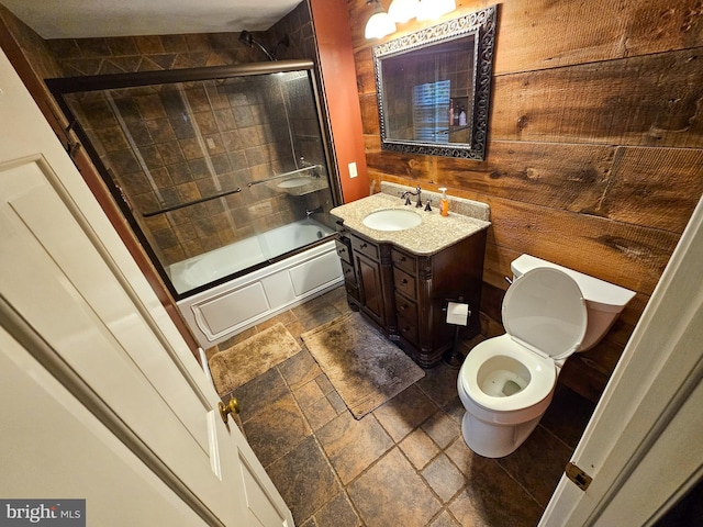 bathroom with stone tile flooring, wooden walls, vanity, toilet, and combined bath / shower with glass door