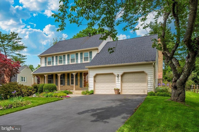 colonial-style house featuring driveway, an attached garage, covered porch, a front yard, and a shingled roof