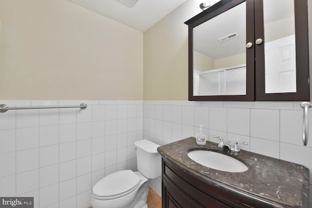 bathroom with vanity, toilet, a wainscoted wall, a shower with door, and tile walls
