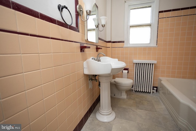 bathroom with radiator, a tub to relax in, tile walls, toilet, and wainscoting