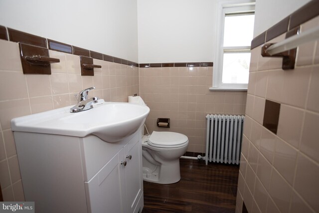 half bathroom with radiator heating unit, tile walls, wood finished floors, vanity, and toilet