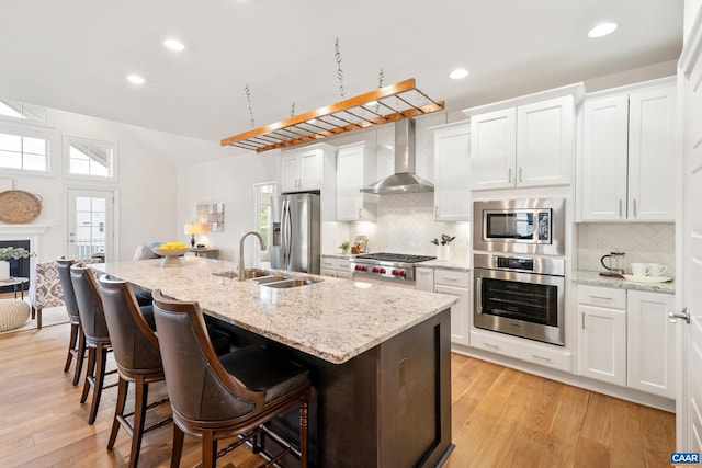 kitchen featuring stainless steel appliances, wall chimney exhaust hood, a sink, a kitchen island with sink, and decorative backsplash