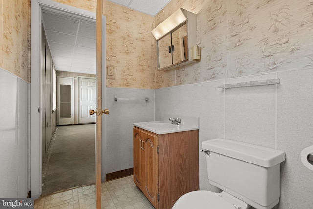 half bathroom featuring wainscoting, toilet, wallpapered walls, vanity, and tile patterned floors