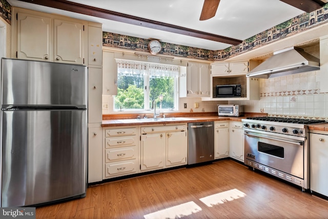 kitchen with stainless steel appliances, wall chimney range hood, a sink, backsplash, and light wood-style floors