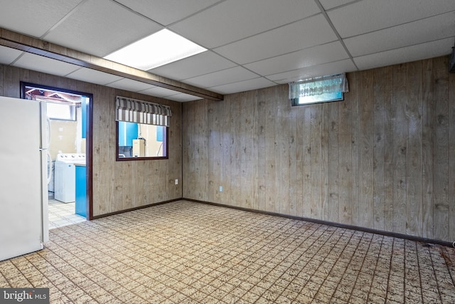 below grade area featuring freestanding refrigerator, a drop ceiling, washer / clothes dryer, wood walls, and plenty of natural light