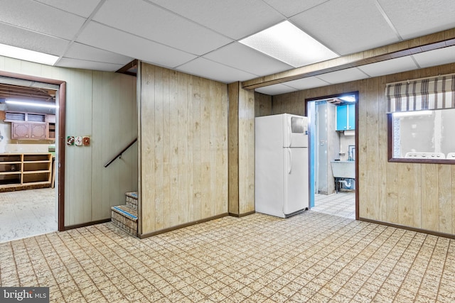 basement featuring freestanding refrigerator, wood walls, a drop ceiling, tile patterned floors, and baseboards