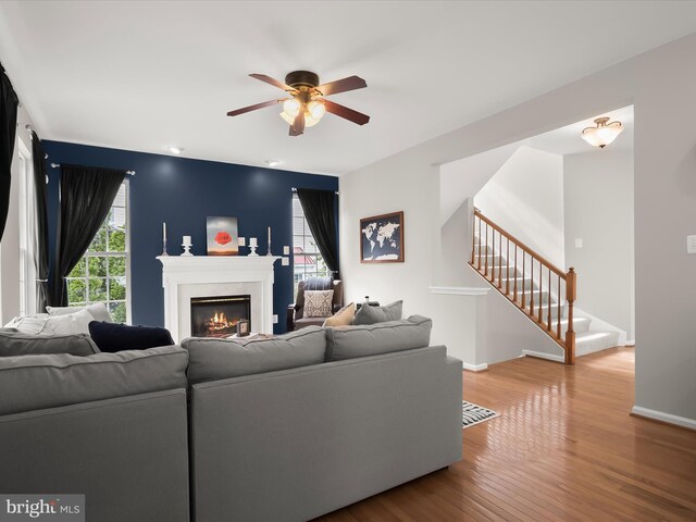 living room featuring stairs, a ceiling fan, a warm lit fireplace, wood finished floors, and baseboards
