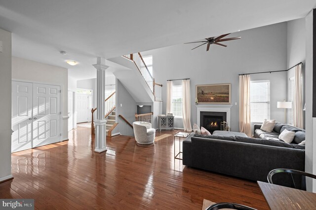 living area featuring a ceiling fan, stairway, a warm lit fireplace, hardwood / wood-style flooring, and a towering ceiling