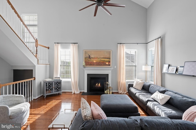 living area with high vaulted ceiling, a ceiling fan, wood finished floors, a fireplace with flush hearth, and stairway