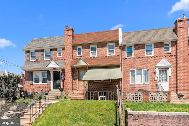 view of property featuring brick siding, a chimney, a front yard, a shingled roof, and entry steps