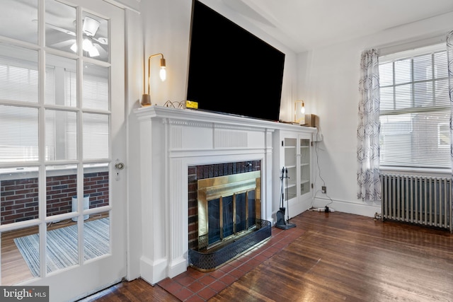 living area featuring radiator, a glass covered fireplace, wood finished floors, baseboards, and a ceiling fan