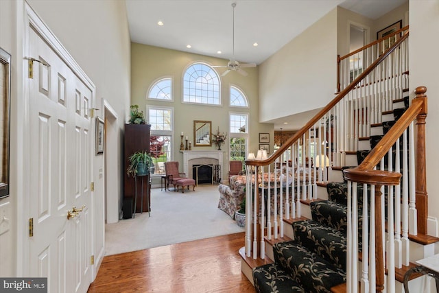 carpeted foyer featuring stairway, a ceiling fan, a glass covered fireplace, a high ceiling, and wood finished floors