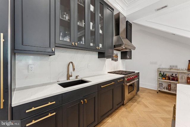 kitchen featuring a sink, range with two ovens, wall chimney range hood, glass insert cabinets, and tasteful backsplash