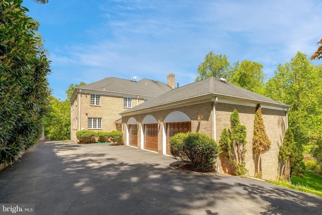 view of property exterior featuring driveway, brick siding, an attached garage, a chimney, and a shingled roof