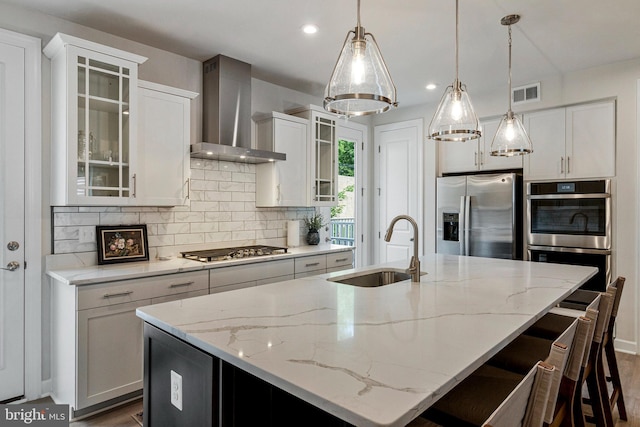 kitchen with a sink, stainless steel appliances, wall chimney exhaust hood, a kitchen island with sink, and tasteful backsplash