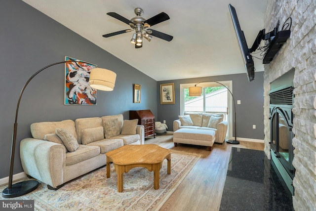 living area with vaulted ceiling, wood finished floors, a ceiling fan, a stone fireplace, and baseboards
