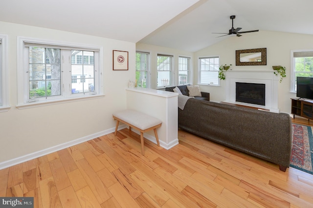 living area with a glass covered fireplace, light wood finished floors, baseboards, a ceiling fan, and vaulted ceiling