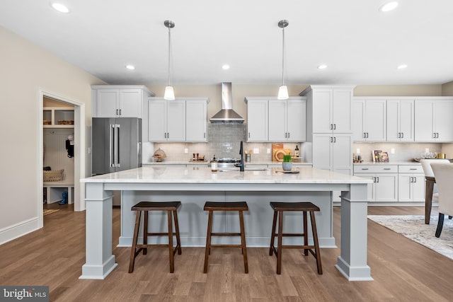 kitchen with wall chimney range hood, decorative backsplash, wood finished floors, white cabinets, and a large island with sink