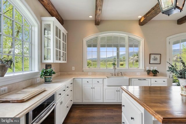 kitchen with a sink, stainless steel oven, beamed ceiling, glass insert cabinets, and white cabinetry